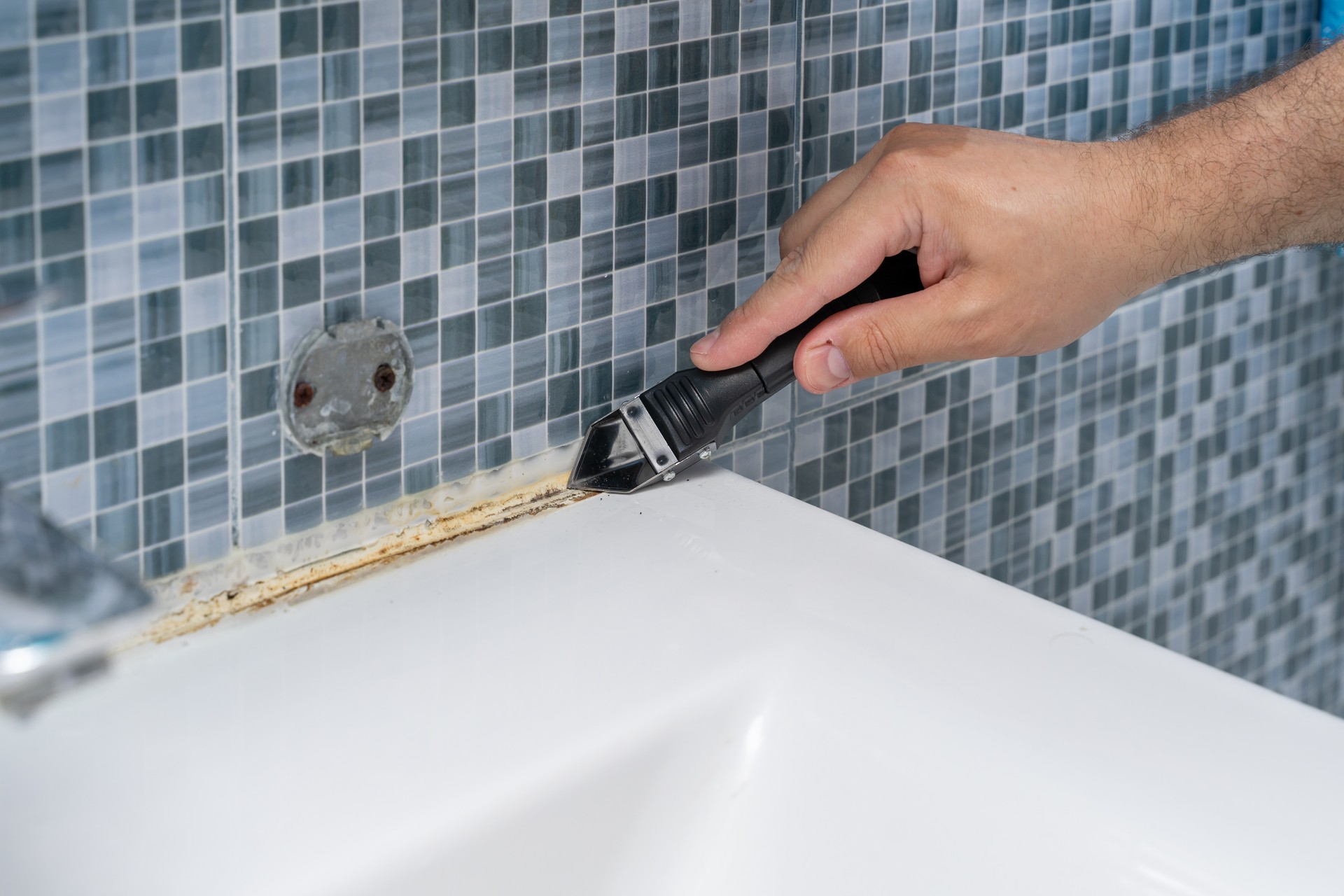 Plumber removes old dirty silicone from tub, cuts silicone adhesive with cutter