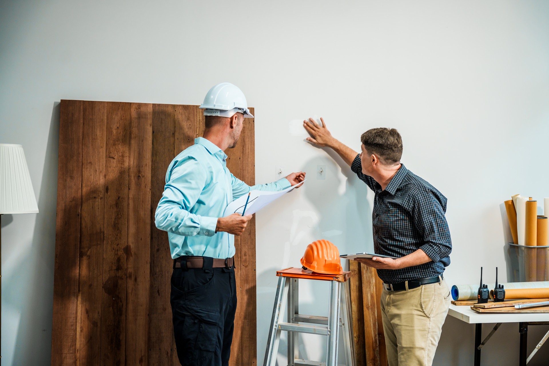 A Caucasian male engineer and an Italian male building contractor are planning and talking at a desk, reviewing house plans and a model of a house with wooden textures for renovation.